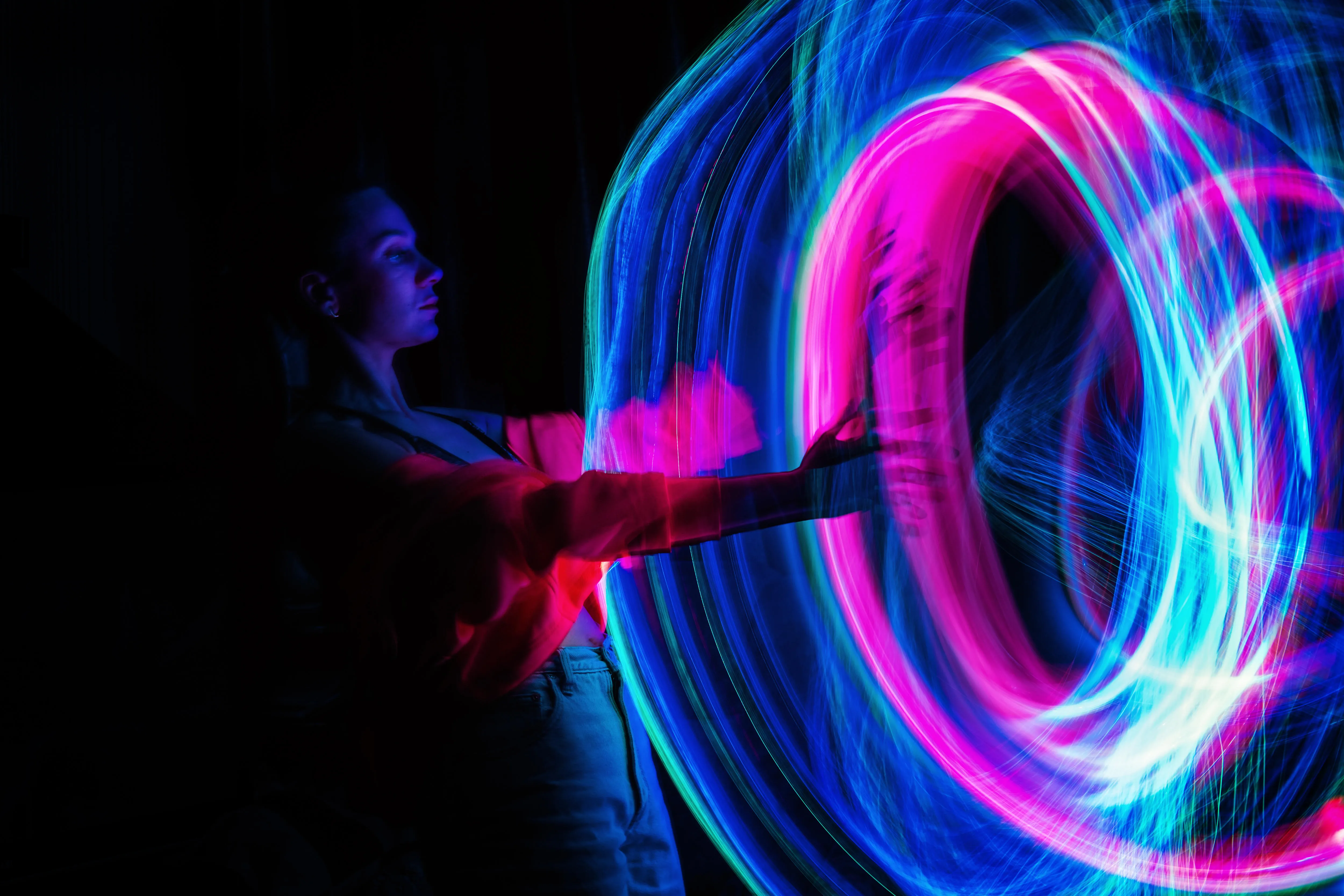 A woman stands in the dark embracing a swirl of vibrant neon colours in shades of pink and blue.