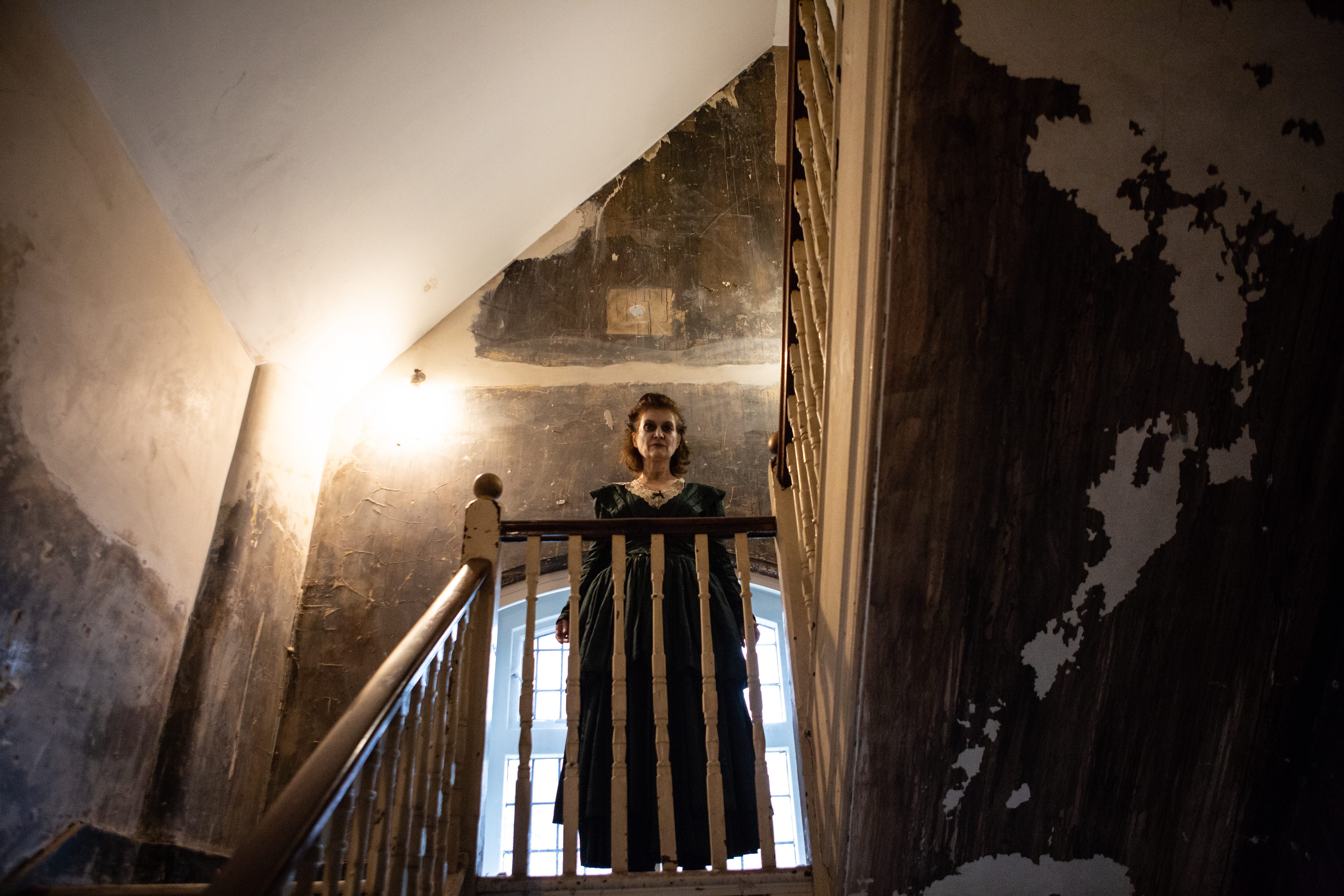 A lady with ghostly makeup in a black dress, poised and standing on a flight of stairs.